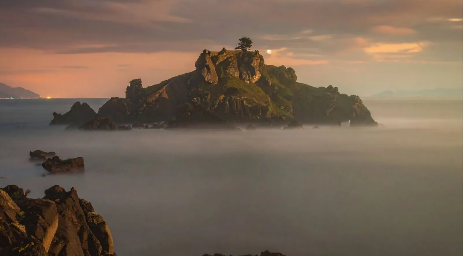 Gaztelugatxe islet silhouetted at dusk with a long-exposure sea and a rising moon behind the rock