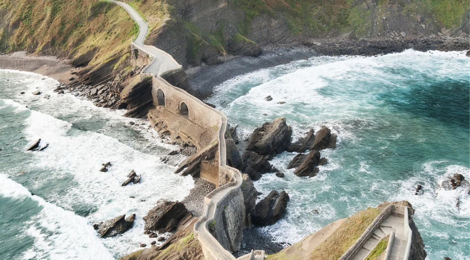 Close aerial view of the arched stone bridge and the first flights of steps at Gaztelugatxe, with surf breaking either side