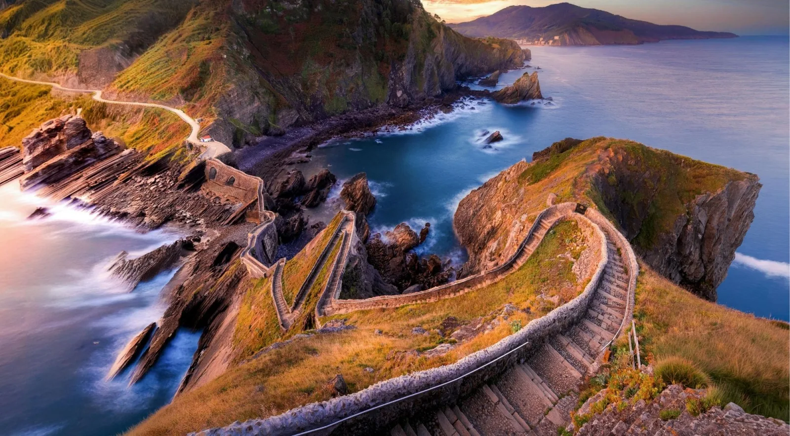 Golden-hour view down the switchback stone staircase of Gaztelugatxe toward the cove and the Basque coastline beyond