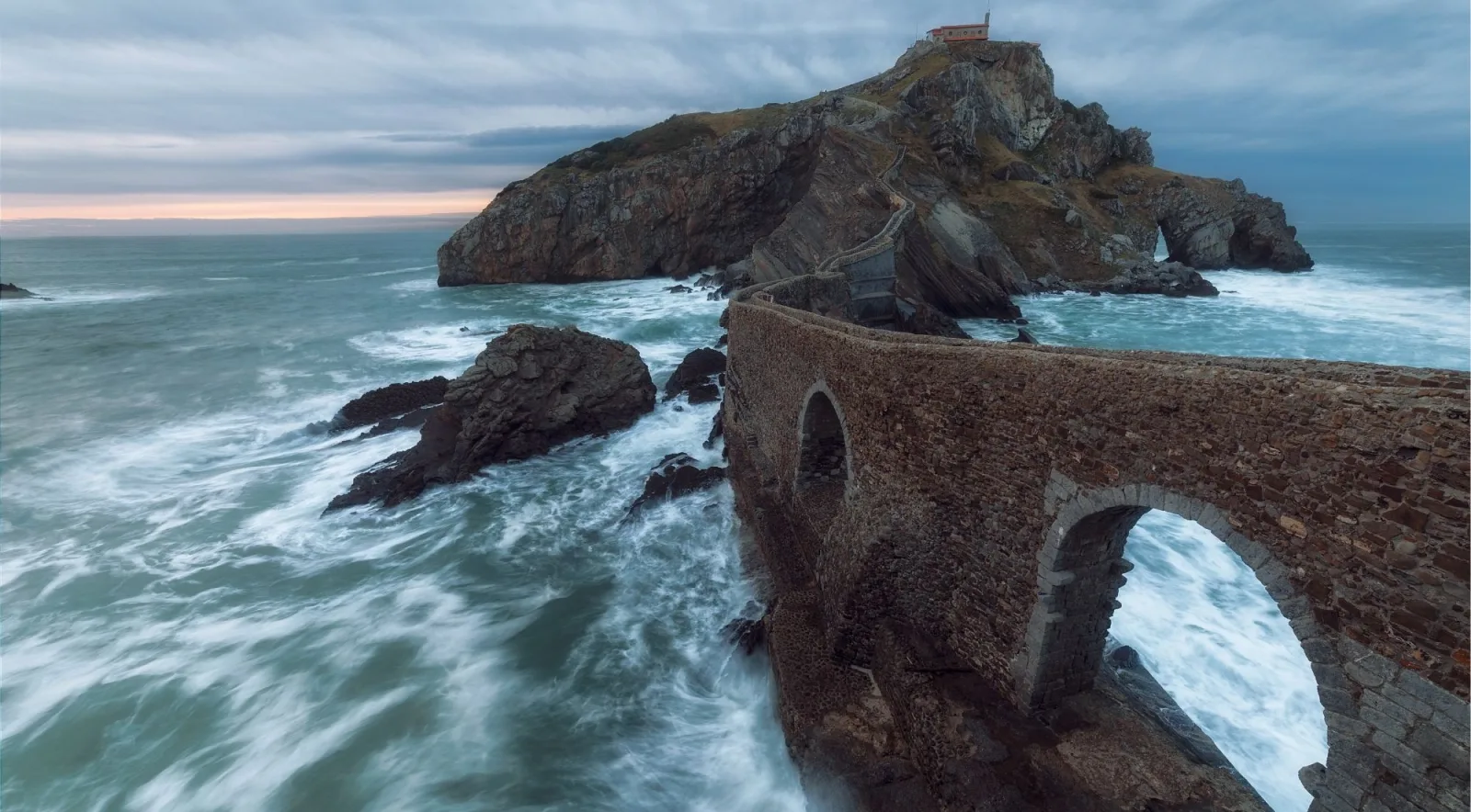 Stormy dusk at Gaztelugatxe with waves washing around the arched stone bridge and the hermitage on the summit