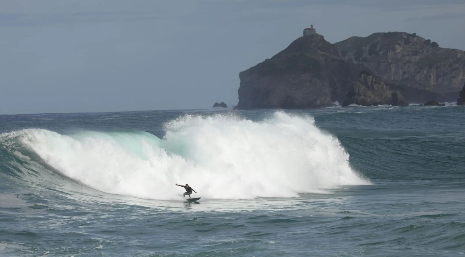 A surfer riding a breaking wave off the Basque coast with the Gaztelugatxe headland in the background