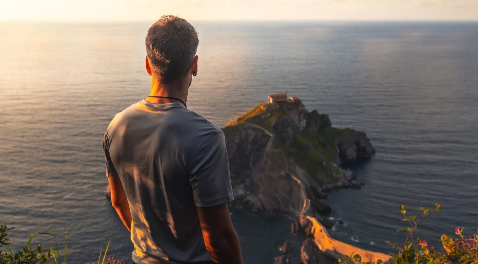 A visitor standing at a clifftop viewpoint at sunset looking across to the Gaztelugatxe hermitage