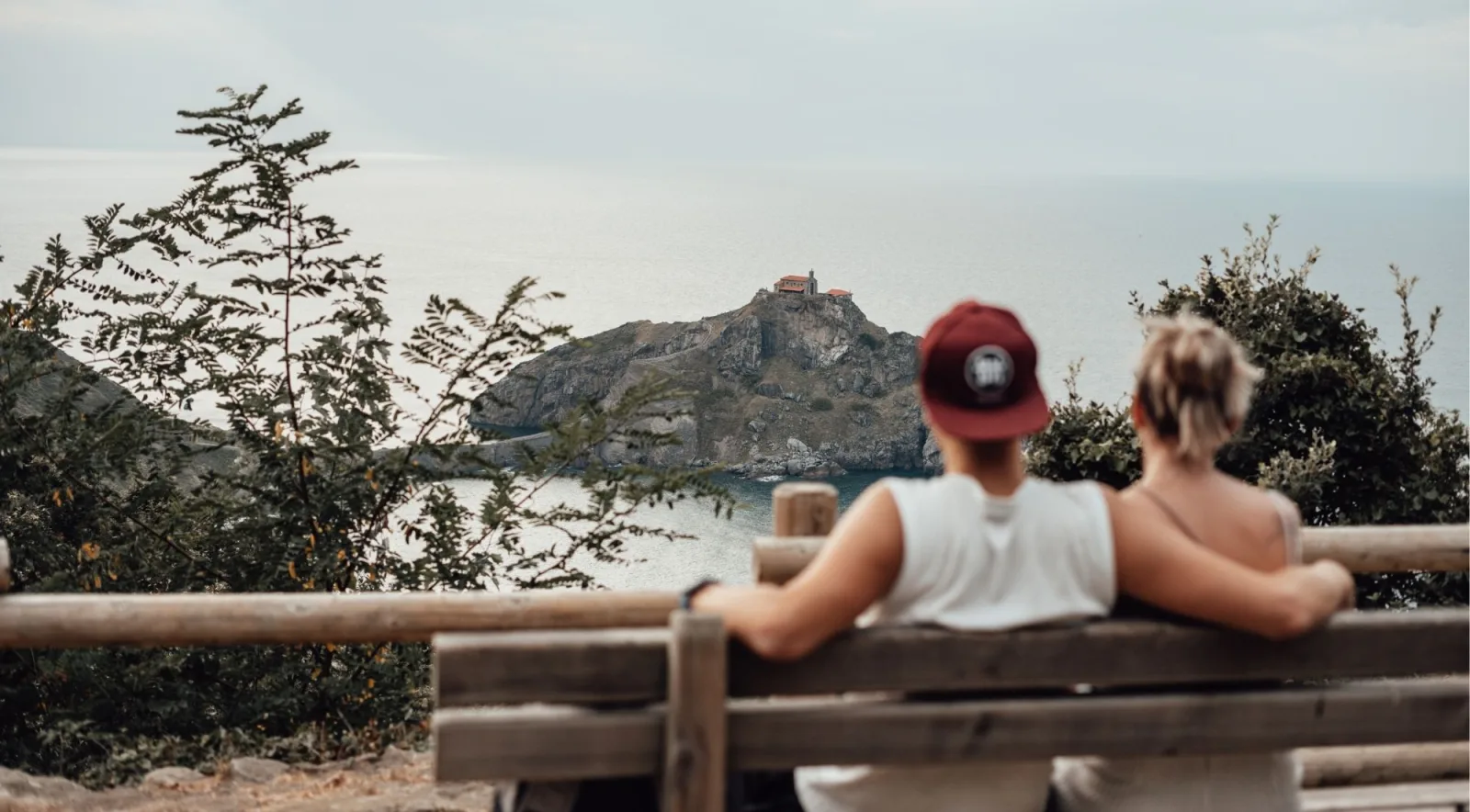 Two travellers sitting on a wooden bench at a viewpoint with Gaztelugatxe framed between the trees